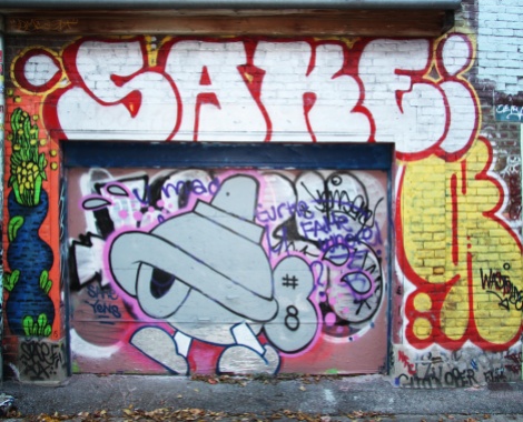 Turtle Caps on garage door, with Waxhead on the left, Sake above and Wastoids on the right, in a graffiti alley between St-Laurent and Clark